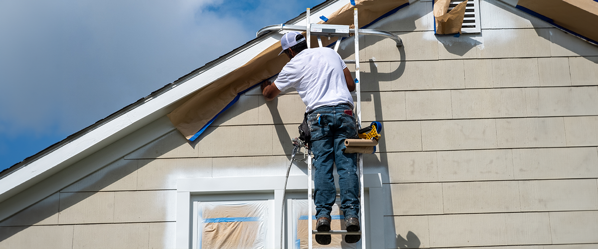 A painter on a ladder working on a house exterior, with masking paper and tape protecting the trim.