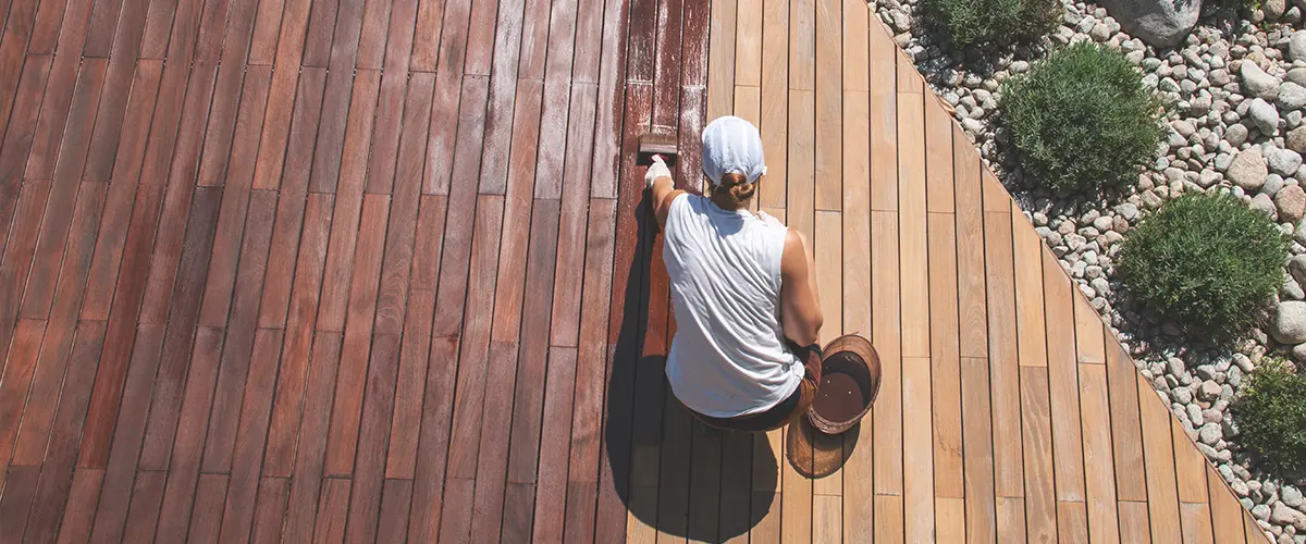 Person applying wood stain to deck for outdoor renovation, showcasing rich color contrast and garden pebbles.