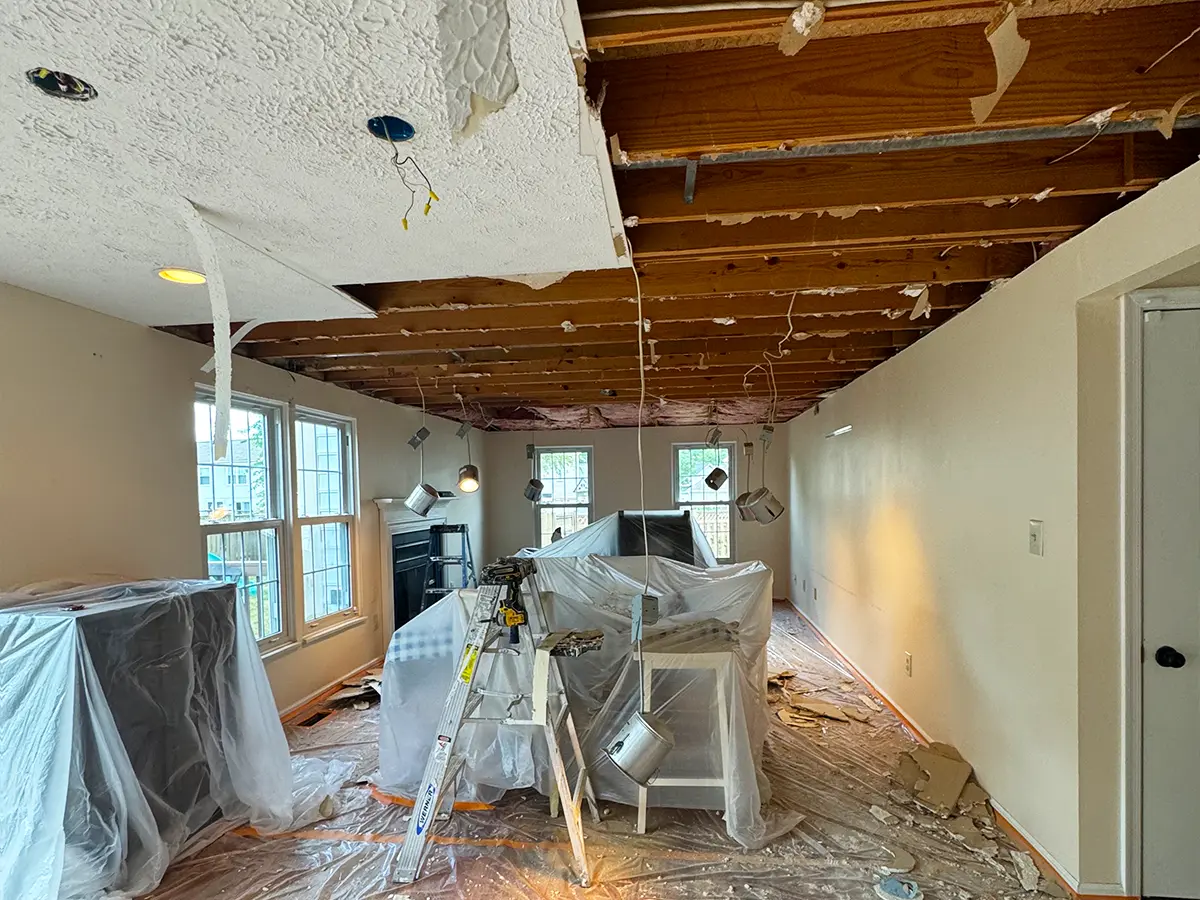 Room during ceiling renovation with exposed beams and covered furniture for drywall replacement preparation.