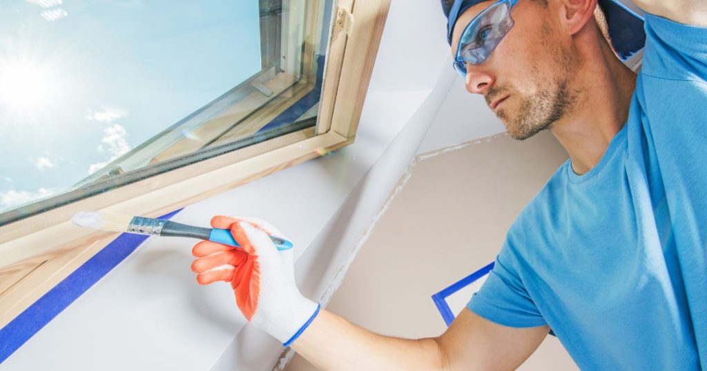 A professional painter wearing protective glasses and gloves carefully painting the trim around a skylight with a brush, ensuring clean edges with blue painter’s tape.