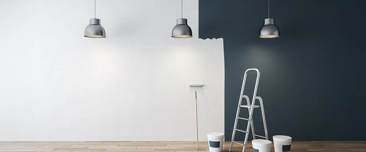 Modern room painting in progress with blue accent wall, metal pendant lights, ladder, and paint buckets on hardwood floor.