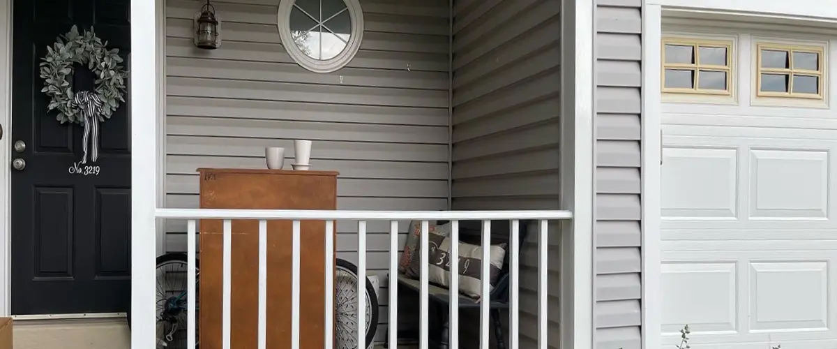 Small front porch with gray vinyl siding, black entry door, white railing, and cozy seating area.
