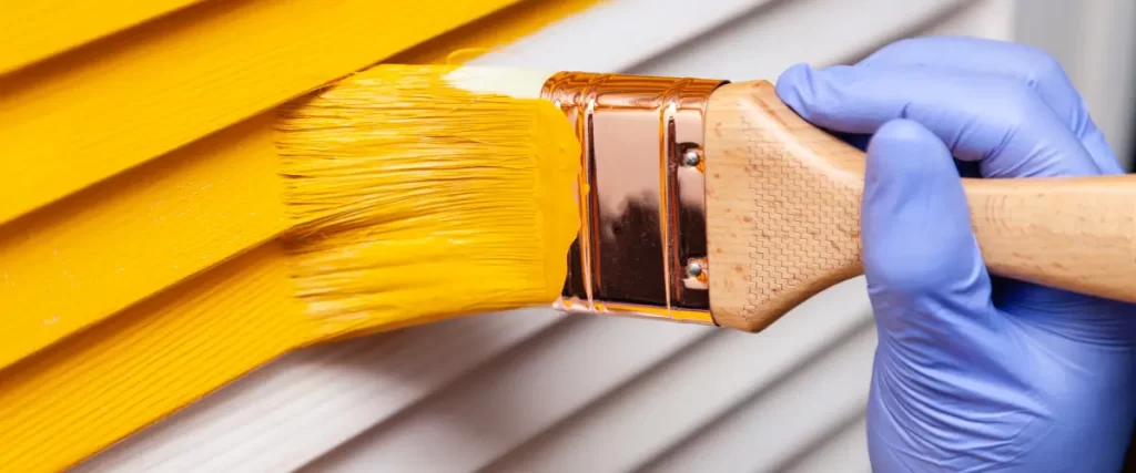 Another close-up of a paintbrush applying yellow paint to siding