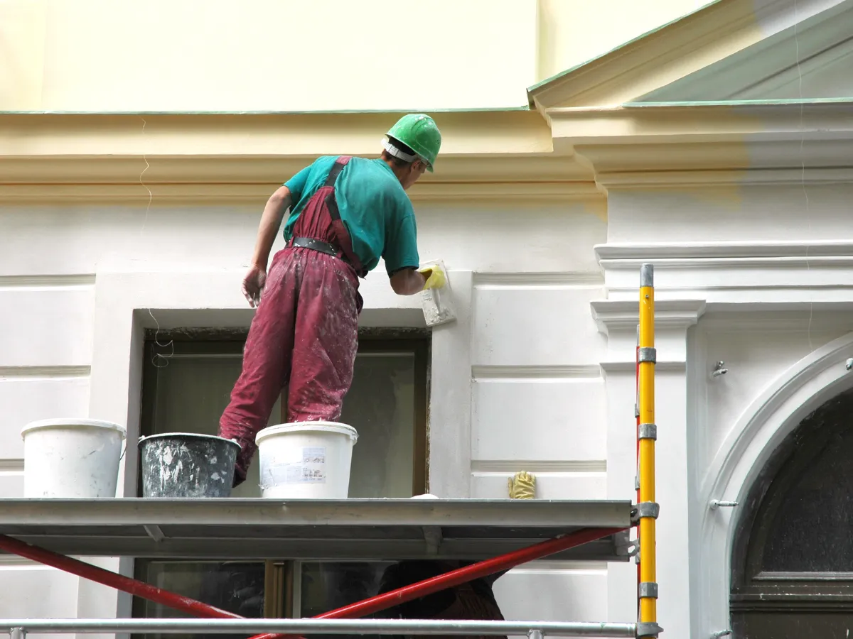 Painter on scaffold working on ornate exterior molding of a light-colored building