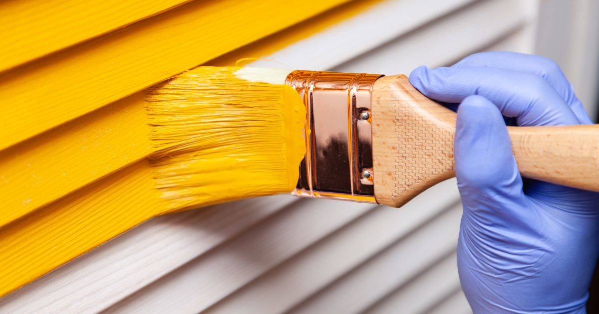 Close-up of a paintbrush applying yellow paint to siding