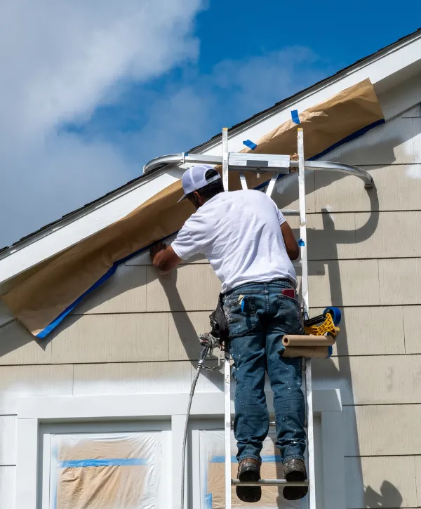 Person painting house trim on a ladder under blue sky