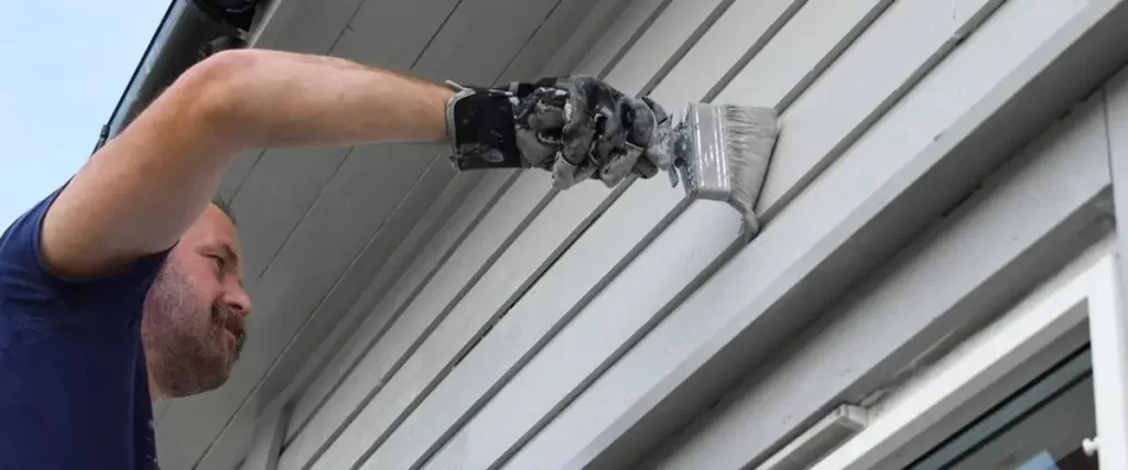 Skilled painter using a brush to apply white paint to exterior siding, showing potential cold weather painting.