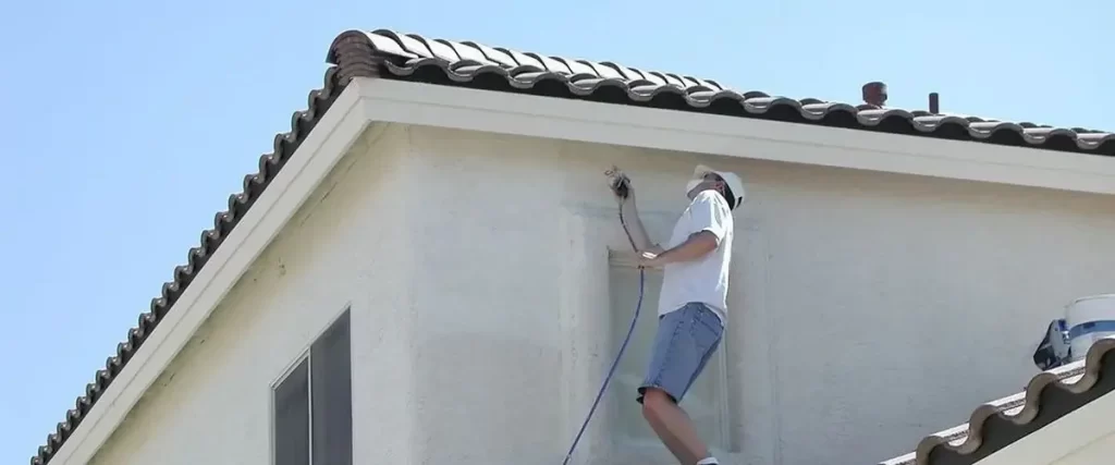 Painter on a ladder using a spray gun to apply paint to the upper level of a stucco house with a tile roof.
